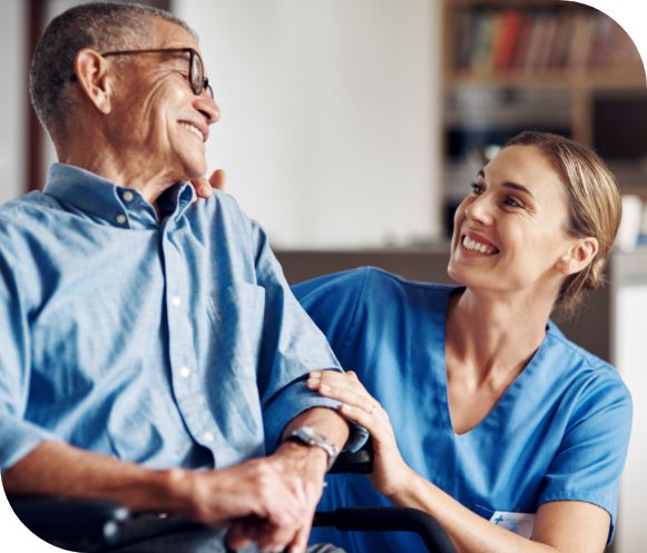 A nurse in blue scrubs kneels beside and smiles at an elderly patient.
